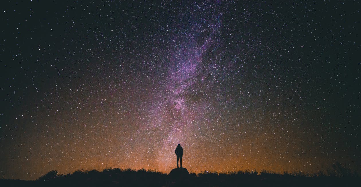 A person standing on a rock under a night sky filled with stars and a visible band of the Milky Way, with a soft orange glow near the horizon.