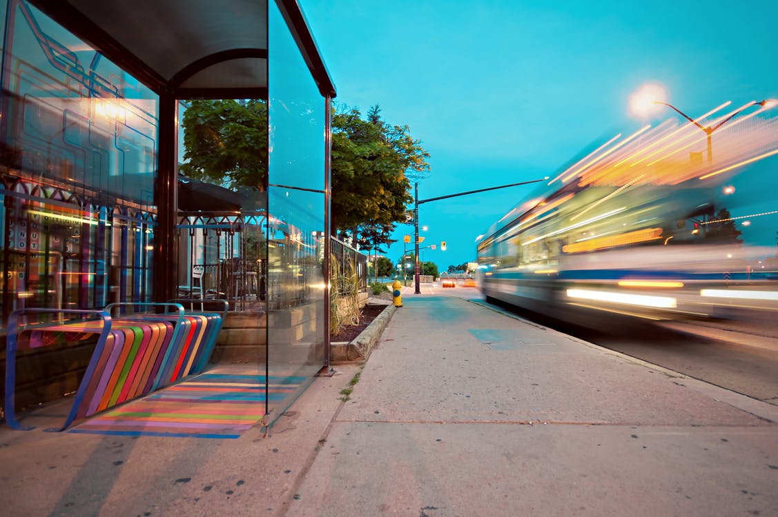 A street scene showing a modern bus shelter with colourful benches and glass panels, as a bus passes by at speed, creating a motion blur under a bright evening sky.